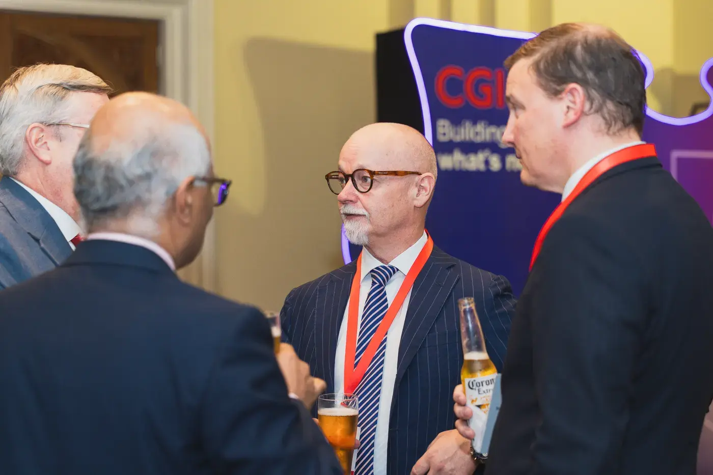 Delegates in suits in conversation, drinks in hand, in front of a lit brand backdrop