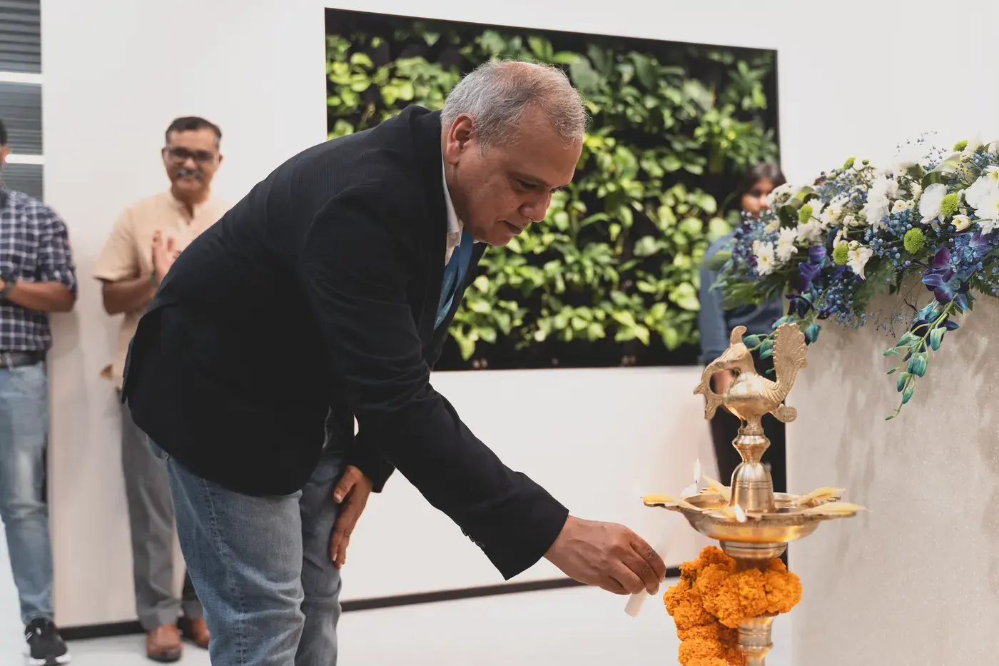 A guest lighting the ceremonial lamp beside a garlanded green wall